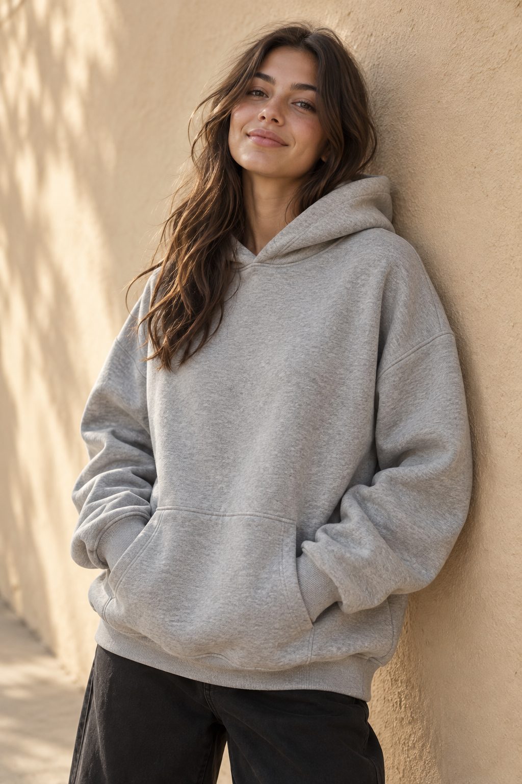 Woman in a heather grey hoodie leaning on a sunlit wall